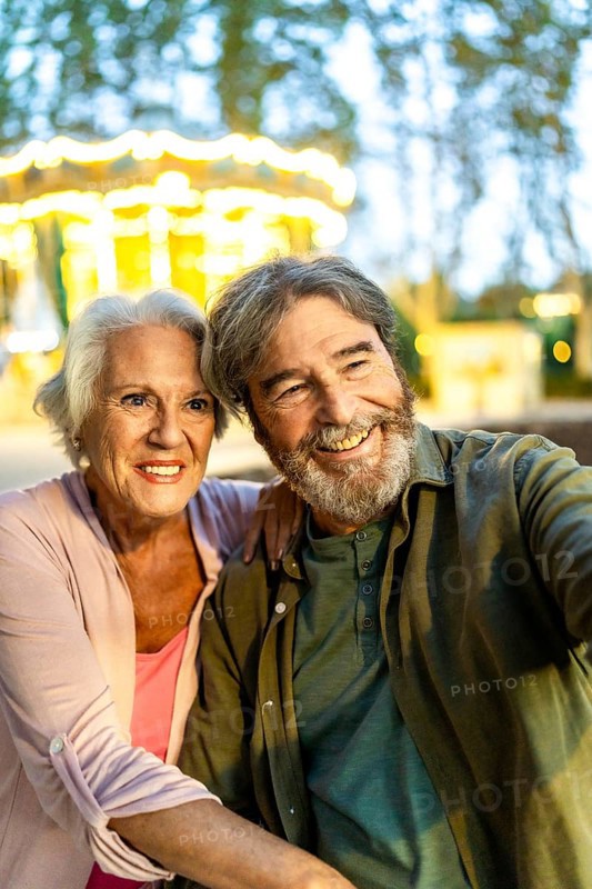 Senior couple taking selfie sitting on bench in the city at night next to carousel