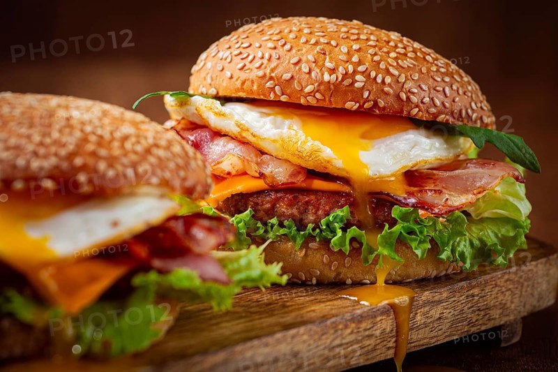 Two juicy burgers sit on a wooden board, each filled with lettuce, bacon, cheese, and fried eggs