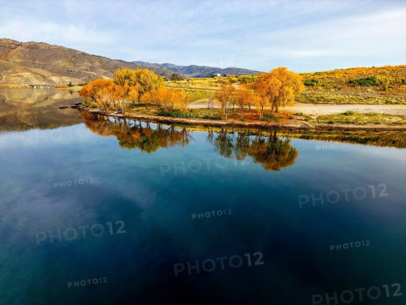 Aerial view of a tranquil lake in Queenstown during autumn, showcasing vibrant foliage and clear reflections on the water surface. Captured by a drone for a stunning aerial perspective