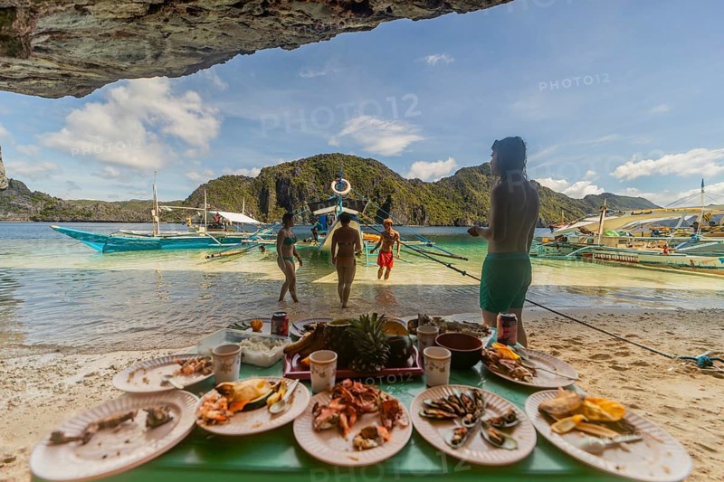 Unrecognizable tourists enjoy a seaside meal in the Philippines, showcasing a vibrant beach background with traditional boats and lush mountains