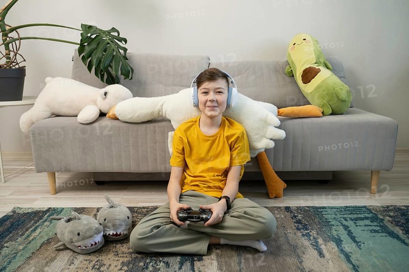 A cheerful boy in a yellow shirt and headphones plays video games, seated in a cozy room with plush toys and a monstera plant on a comfy sofa