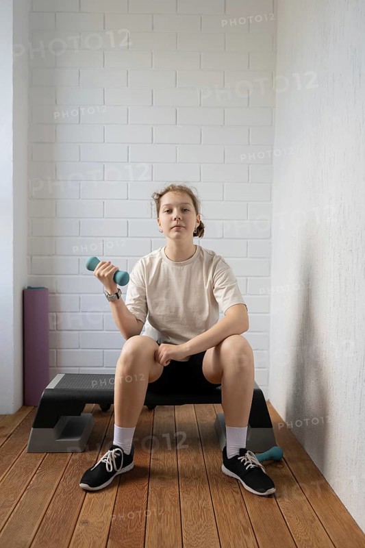 A teen girl confidently exercises with a dumbbell in a home gym setting, showcasing her commitment to fitness and healthy lifestyle on a wooden floor with gym equipment nearby
