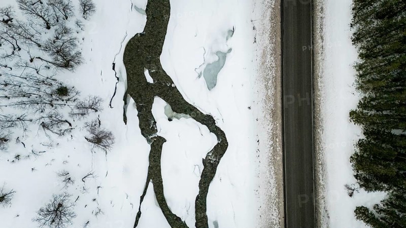 Aerial view of a Nordic landscape showcasing a road flanked by a frozen stream and snow-laden trees. The serene winter scene captures nature's tranquil beauty, Bardu, Troms, Norway
