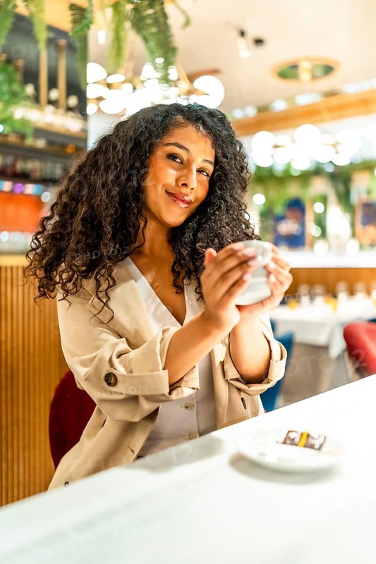 Vertical portrait of a latin beauty woman enjoying warm coffee sitting in a modern cafeteria smiling at camera