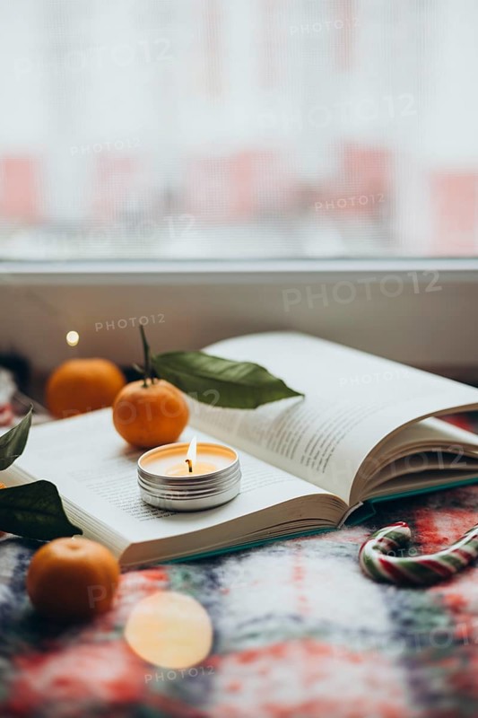 Warm and inviting Christmas scene featuring a lit candle placed on an open book, surrounded by fresh tangerines and a candy cane, offering a sense of holiday cheer and relaxation