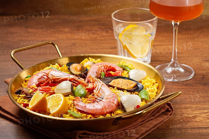 A large dish of seafood paella features shrimp, mussels, and other ingredients. A glass with a drink and slice of lemon is nearby on the wooden table. The meal is ready to eat