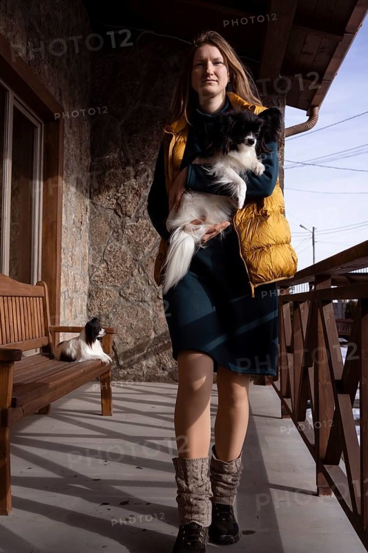A woman stands on a rustic porch holding her purebred papillon dog. Natural light illuminates the charming scene, capturing the bond between owner and pet