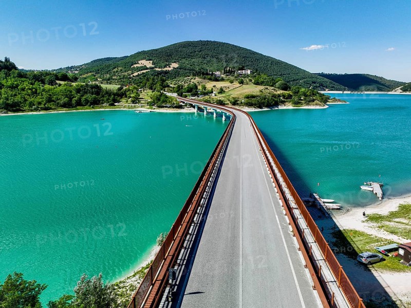 A breathtaking view of a bridge extending over a turquoise lake surrounded by lush hills in Italy, capturing the picturesque charm and serene beauty of Italian nature