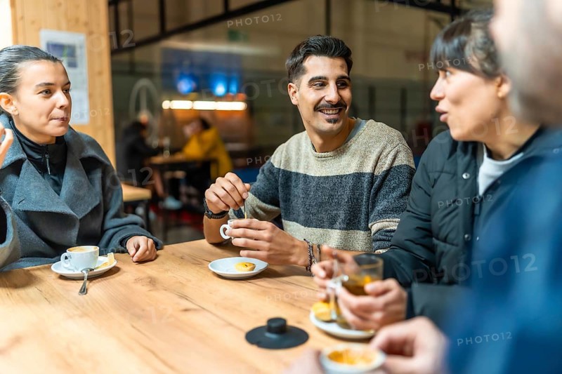 Diverse group of friends socializes and talks while drinking coffee and tea at a wooden table in an indoor cafe setting, building community after a pickleball game