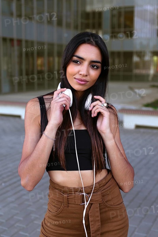 A young woman with long dark hair holds white headphones around her neck while standing outside. She is wearing a black top and brown pants, enjoying a moment of leisure
