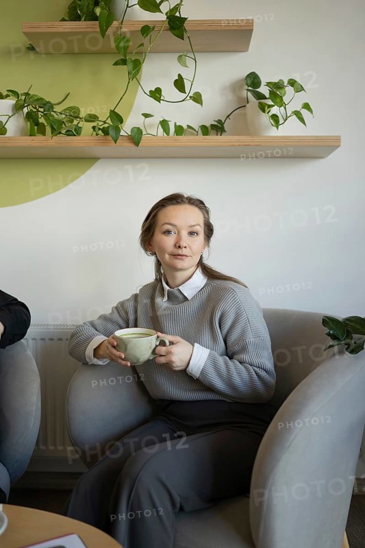 A woman sits comfortably in a cafe, sipping coffee. She is surrounded by green plants, creating a relaxing and inviting atmosphere. The serene scene embodies comfort