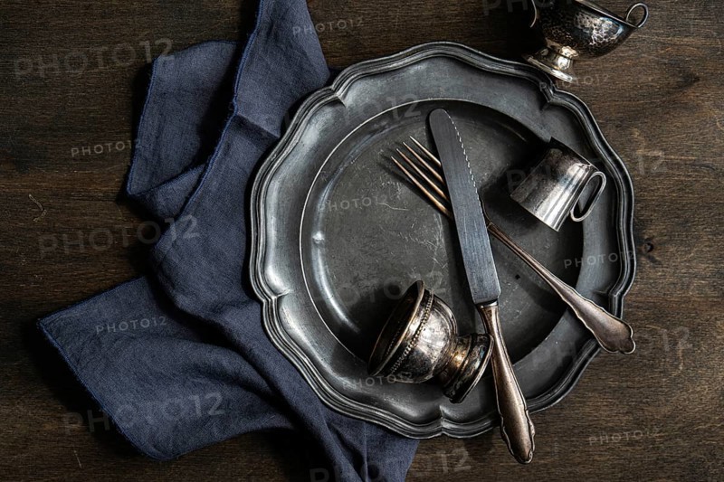 A collection of vintage silver plate, cutlery, and cups arranged on a dark wooden surface with a blue cloth. The tarnished metal adds a rustic, antique touch