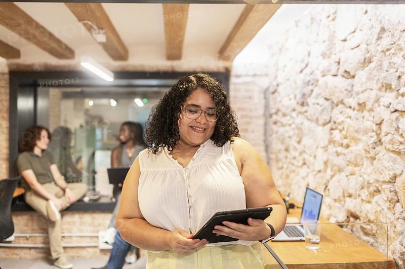 Multiracial coworkers collaborate in a modern office. A woman uses a tablet while others engage in conversation, showcasing a diverse and inclusive work environment