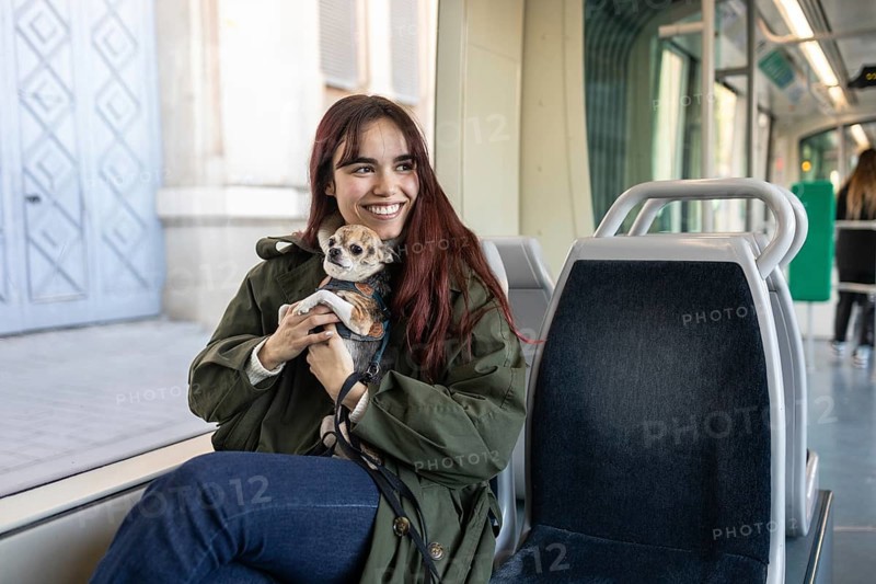 A cheerful woman sits on a tram seat holding her small chihuahua, warmly wrapped in a coat. The tram modern interior creates a cozy setting for their journey