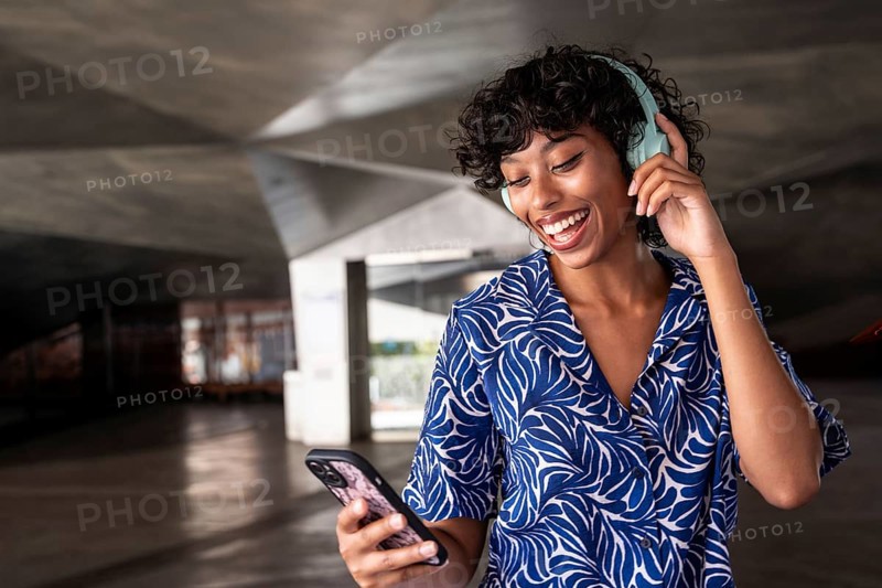 A joyful woman enjoys music through her headphones while holding a smartphone, standing in a modern architectural space. She smiles brightly, expressing contentment