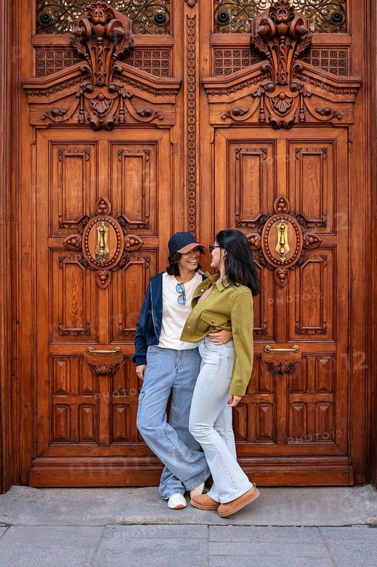 A lesbian couple smiling and embracing in front of an ornate wooden door, exuding love and happiness in an urban environment. Casual and warm atmosphere captured in the city