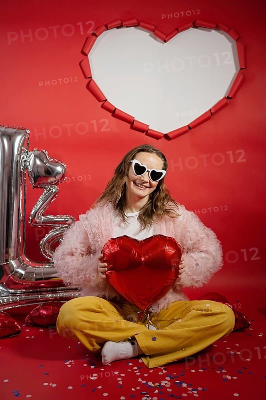 A young woman smiles joyously while holding a red heart-shaped balloon. She wears heart-shaped sunglasses and sits against a vibrant red background with Valentine's Day decorations