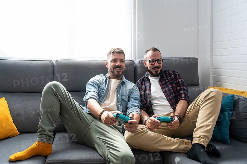 A joyful gay couple relaxes on a couch, engaging in a lively video game. Their cheerful expressions highlight a moment of leisure and connection, capturing modern home life