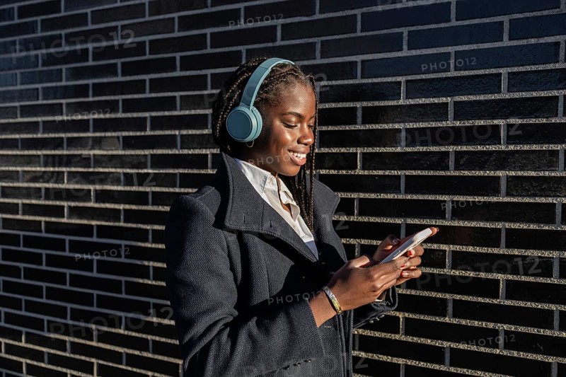 Side view of a young African American businesswoman with braids is seen enjoying a break, smiling while using her smartphone and listening to music through headphones outdoors