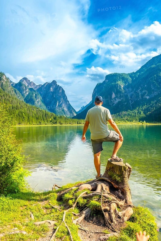 Tourist standing on a tree stump, admiring the stunning landscape of lake dobbiaco and the surrounding dolomites mountains in summer