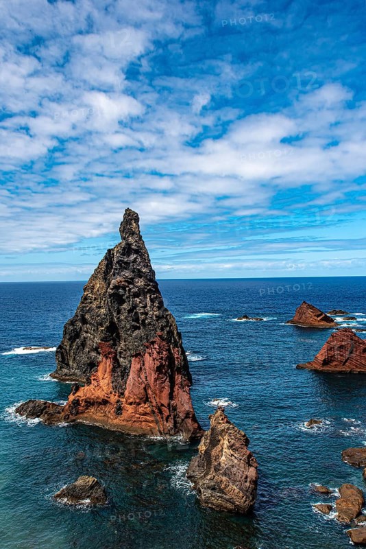 Steep rock formations rise out of the deep blue sea under a partly cloudy sky