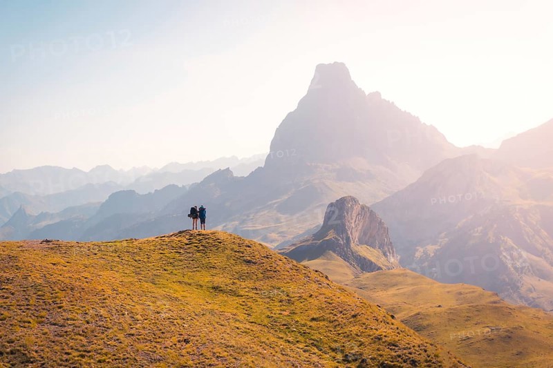 Two hikers stand on a grassy hill, gazing at the breathtaking mountain range in the Pyrenees. The vast landscape and early light create a serene and inspiring scene in summer