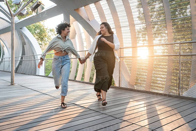 Lesbian couple joyfully holds hands while running together on a modern walkway, surrounded by sunlight and greenery. A moment of freedom and happiness captured