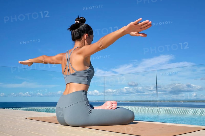 A woman practices yoga by a pool with a stunning ocean view. She sits in lotus pose on a mat, extending her arms with focus and tranquility under a clear blue sky