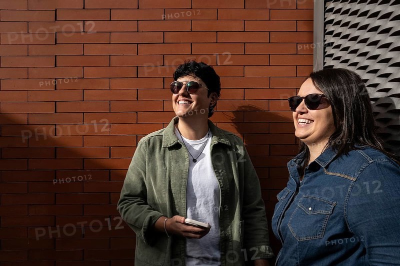 A happy curvy lesbian couple enjoying a sunny day outdoors. They are smiling, wearing casual jackets and sunglasses, embracing love and togetherness against a brick wall backdrop
