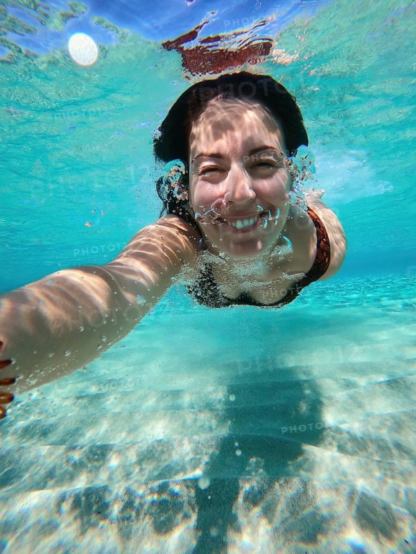 A woman swims underwater in Greece, capturing a selfie. The clear blue water reflects sunlight, creating a playful and joyful atmosphere in the sea