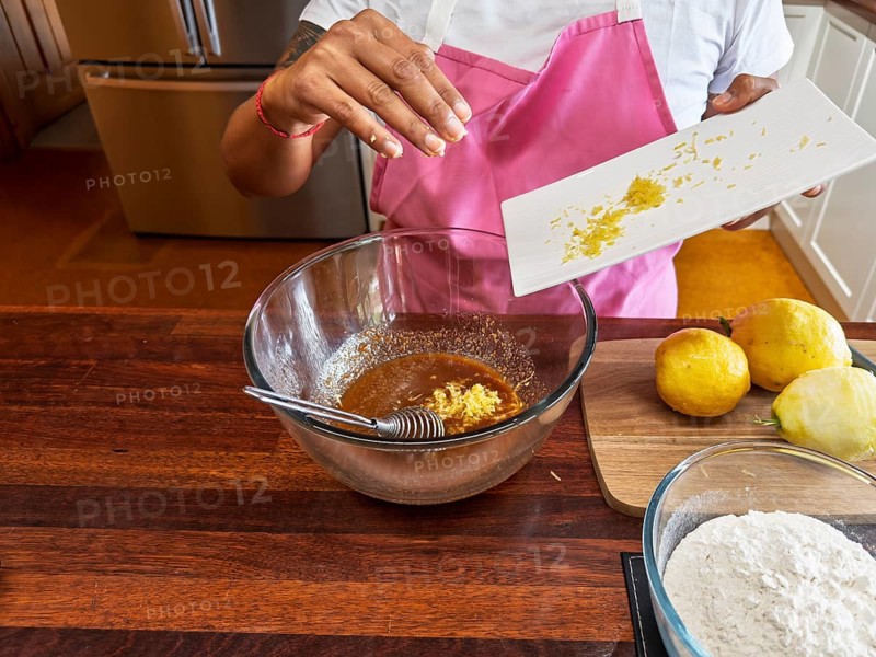 A woman wearing a pink apron prepares lemon cookies in the kitchen. She adds lemon zest to a brown mixture in a glass bowl, with the ingredients arranged on a wooden countertop