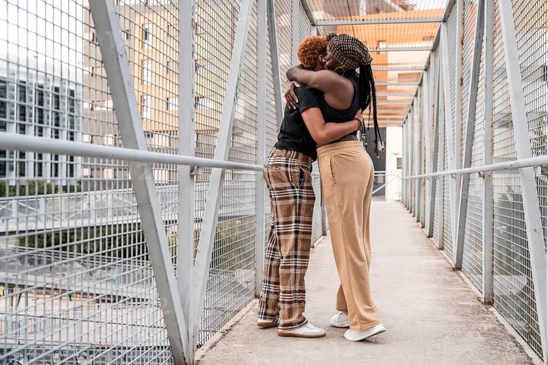 A loving LGBT multiethnic couple embraces on a metal bridge in an urban setting. Their warm hug showcases affection and inclusivity, set against a backdrop of city architecture