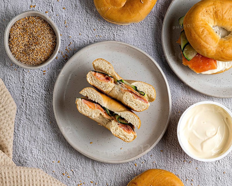 A top view of bagels with salmon and cream cheese on a speckled plate. A bowl of cream cheese and a bowl of seeds are nearby on a textured gray surface