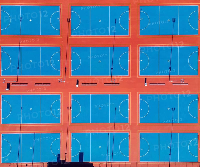 From above, this image captures a series of neatly arranged netball courts in the heart of Canberra, Australia's capital. The drone's perspective highlights the vibrant blue playing surfaces contrasted with bold orange boundaries