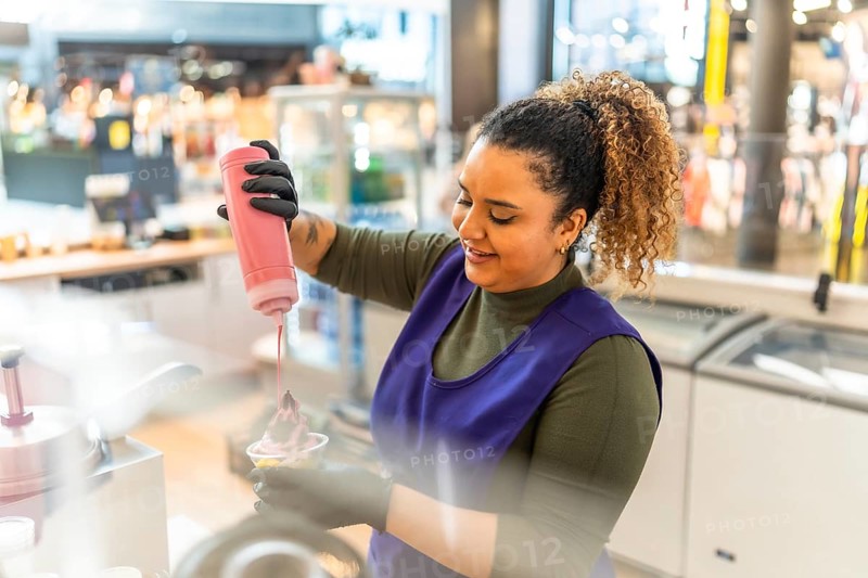 Woman employee in uniform smiling as she pours pink syrup over frozen yogurt in a cup at a shop counter, serving a fresh sweet treat to a customer in a casual small business setting