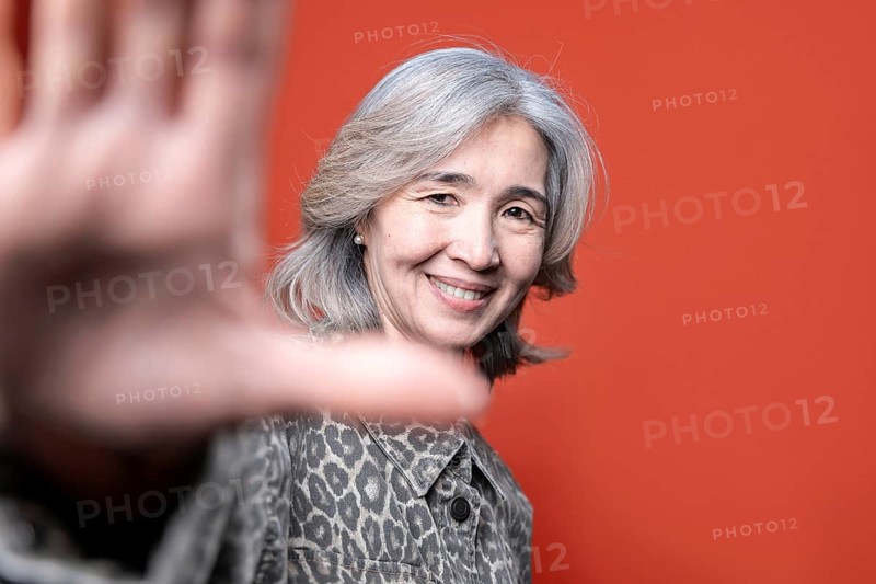 Asian senior woman smiles warmly while posing in front of a vibrant orange background. Her gray hair and patterned jacket add character, conveying confidence and positivity