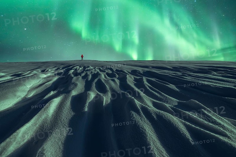 Unrecognizable person in red walks alone under the Northern lights on a snowy landscape at night, showcasing the beauty of a winter northern night
