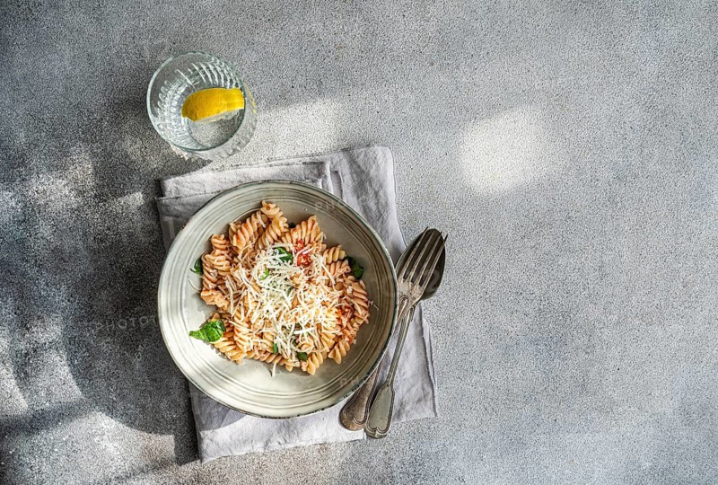 A delectable plate of cavatappi pasta topped with fresh basil, cheese and spicy tomato sauce accompanied by a glass of water with lemon, resting on a folded napkin with utensils