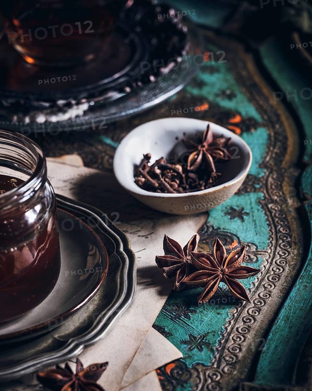 A cozy arrangement featuring a cup of tea infused with anise, presented on an ornate table. The scene includes a glass jar, star anise pods, and a delicate bowl on a vintage surface