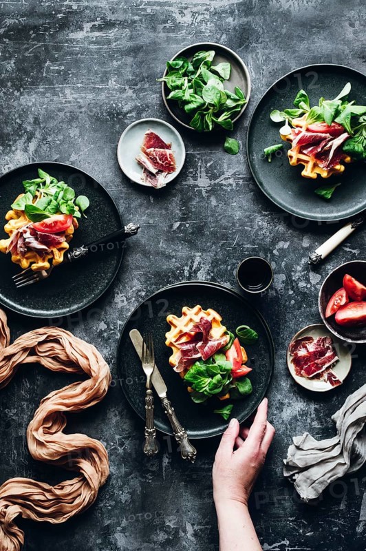 Elegant gourmet dish featuring pumpkin waffles beautifully topped with savory Iberian ham, fresh greens, and ripe tomato slices, creating a delightful balance of flavors