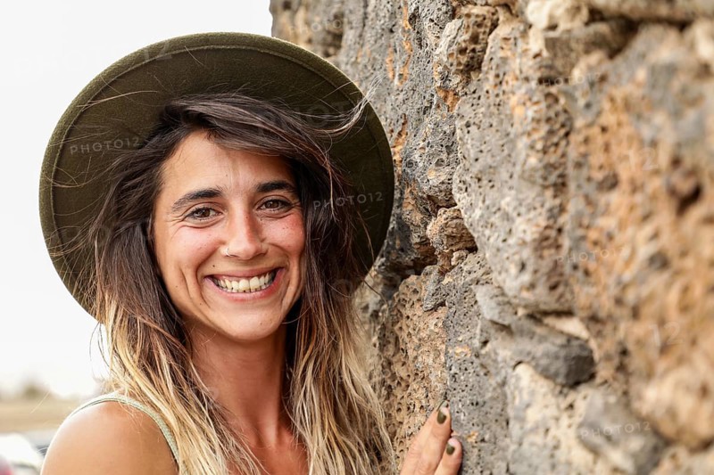 A woman wearing a hat smiles brightly while standing against a textured stone wall Her natural, carefree expression captures the essence of joyful outdoor adventures