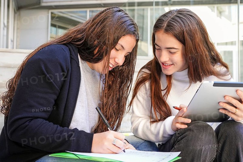 Two students are sitting outdoors, working together on a project One is writing on paper, while the other holds a tablet, indicating teamwork and digital learning