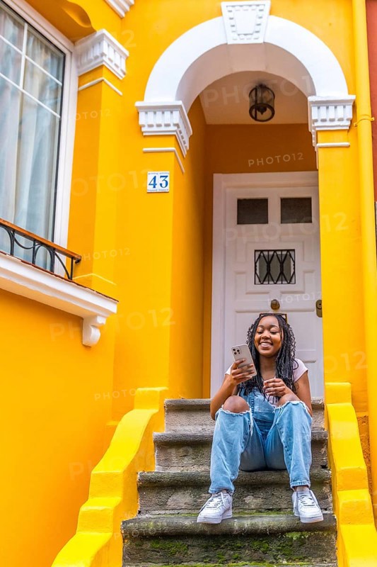 Vertical portrait of a smiling african woman using phone sitting in outside stairs of a yellow colored house
