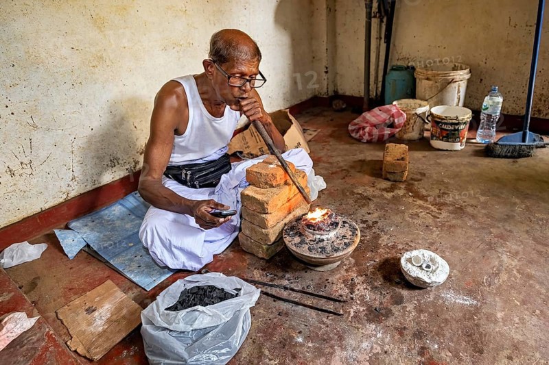 A man sits in a workshop, uses a bellows to start a fire, a man burns sapphires in a small oven in the town of Ratnapura in Sri Lanka