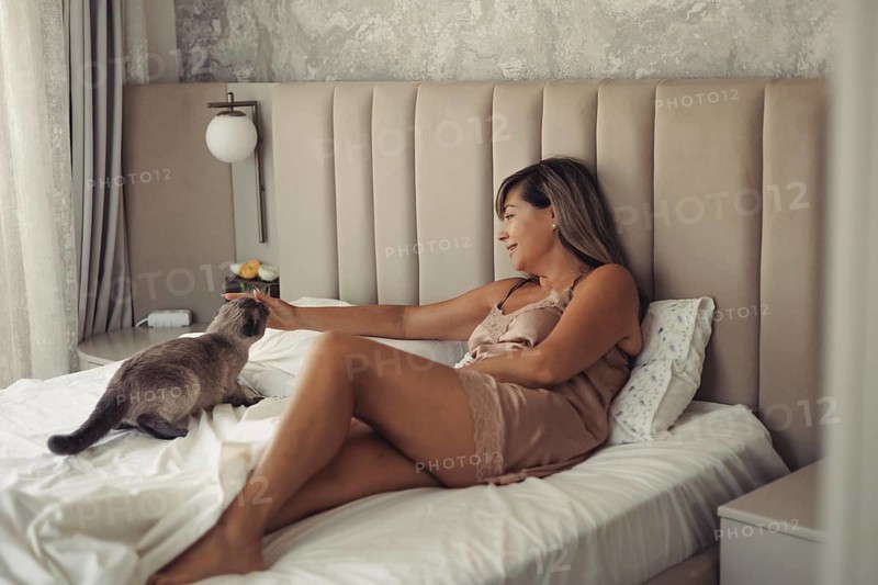 A woman in a satin nightgown relaxes on her bed, gently petting her cat in a cozy, sunlit bedroom. The ambiance exudes comfort, warmth, and the joy of home life