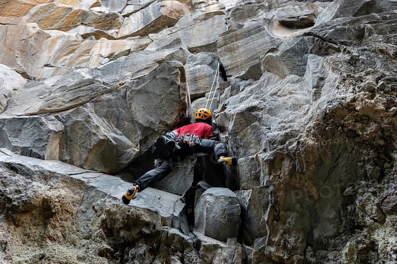 A climber in a red shirt and helmet is scaling a rugged mountain face. The image captures the essence of traditional climbing in nature, emphasizing skill and determination