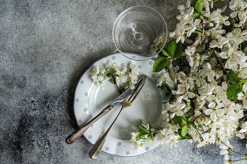 A table setting features vintage cutlery on a patterned plate, adorned with delicate white blossoms. A nearby crystal glass adds a touch of elegance to the rustic scene
