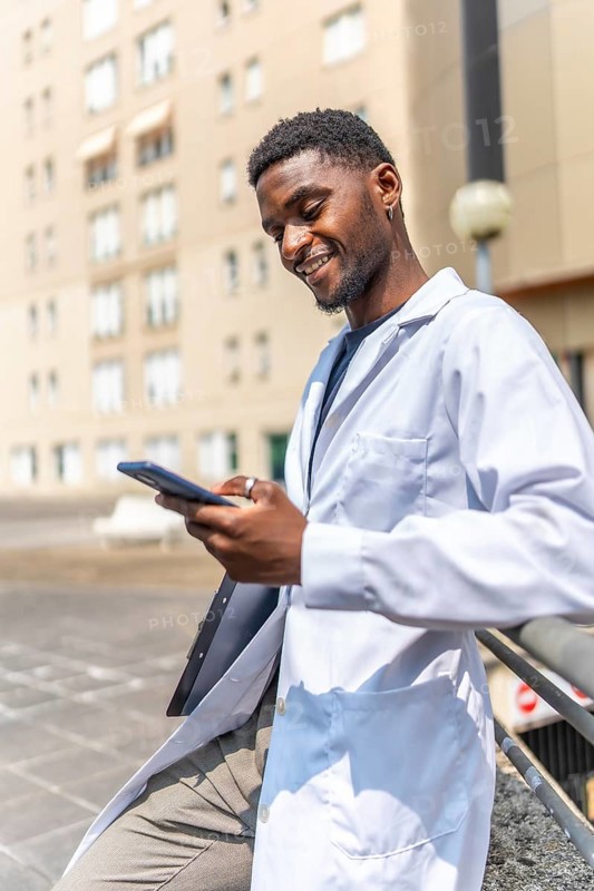 Vertical photo of a cheerful young african doctor using phone during work break outside the hospital