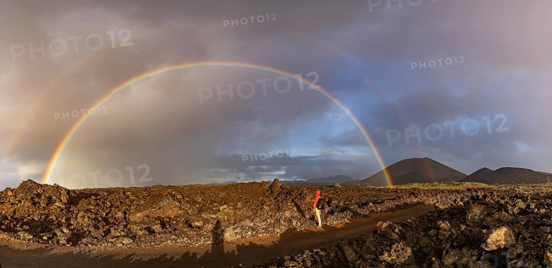 A vibrant rainbow arches over the rugged volcanic terrain of Timanfaya National Park in Lanzarote, Canary Islands, with a solitary figure standing in awe of the natural beauty