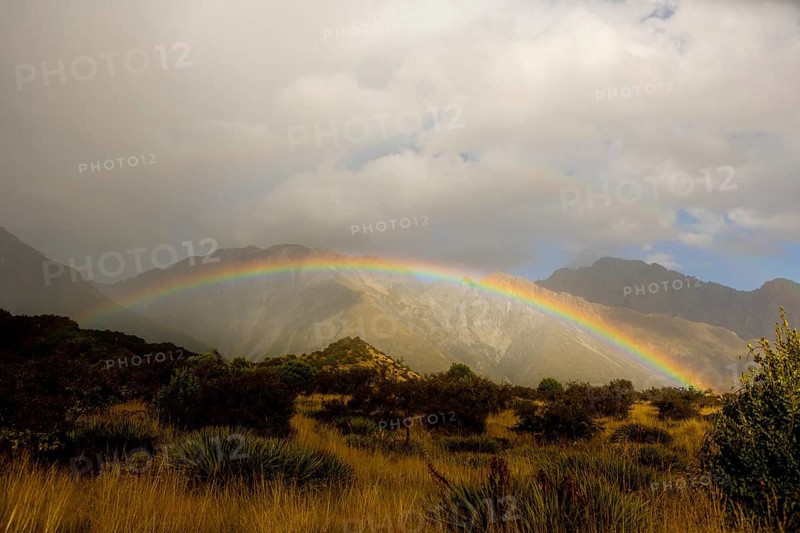 A breathtaking autumn scene at Mount Cook, New Zealand, featuring a vibrant rainbow arching over rugged mountains and lush greenery, under a dramatic cloudy sky